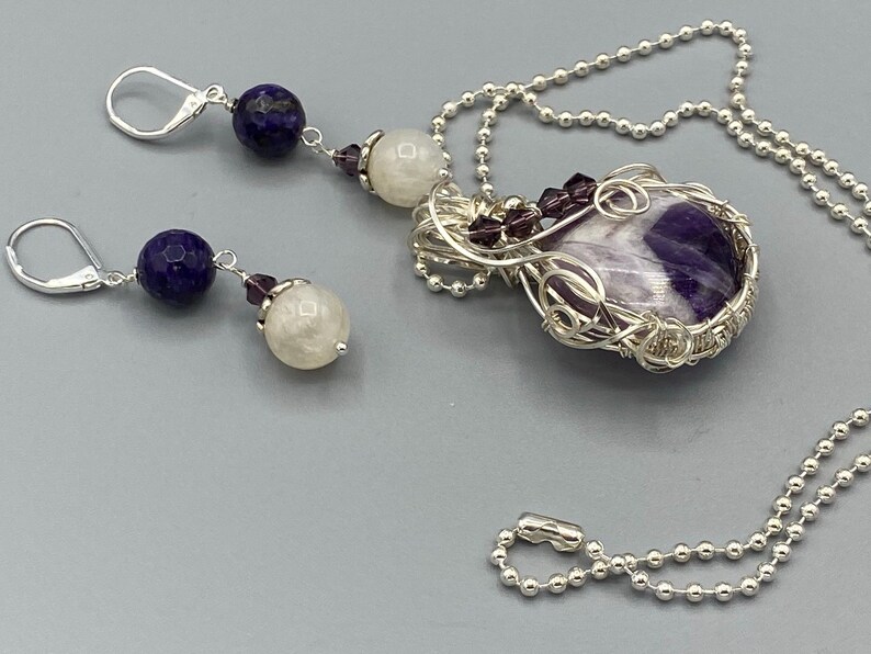 May include: A silver necklace with a purple and white amethyst stone pendant wrapped in silver wire. The necklace has a silver chain and a silver clasp. The pendant is surrounded by small purple beads. The necklace comes with a pair of earrings with a blue stone, a white stone, and a purple bead.