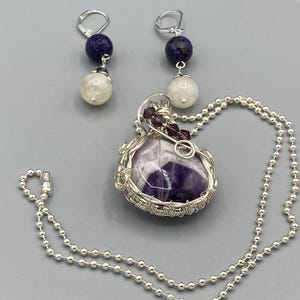 May include: A silver necklace with a purple amethyst pendant wrapped in silver wire. The pendant is accented with small purple crystals. The necklace is paired with a set of earrings featuring a white moonstone and a dark blue stone.