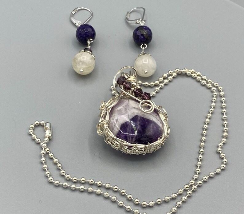 May include: A silver necklace with a purple amethyst pendant wrapped in silver wire. The pendant is accented with small purple crystals. The necklace is paired with a set of earrings featuring a white moonstone and a dark blue stone.