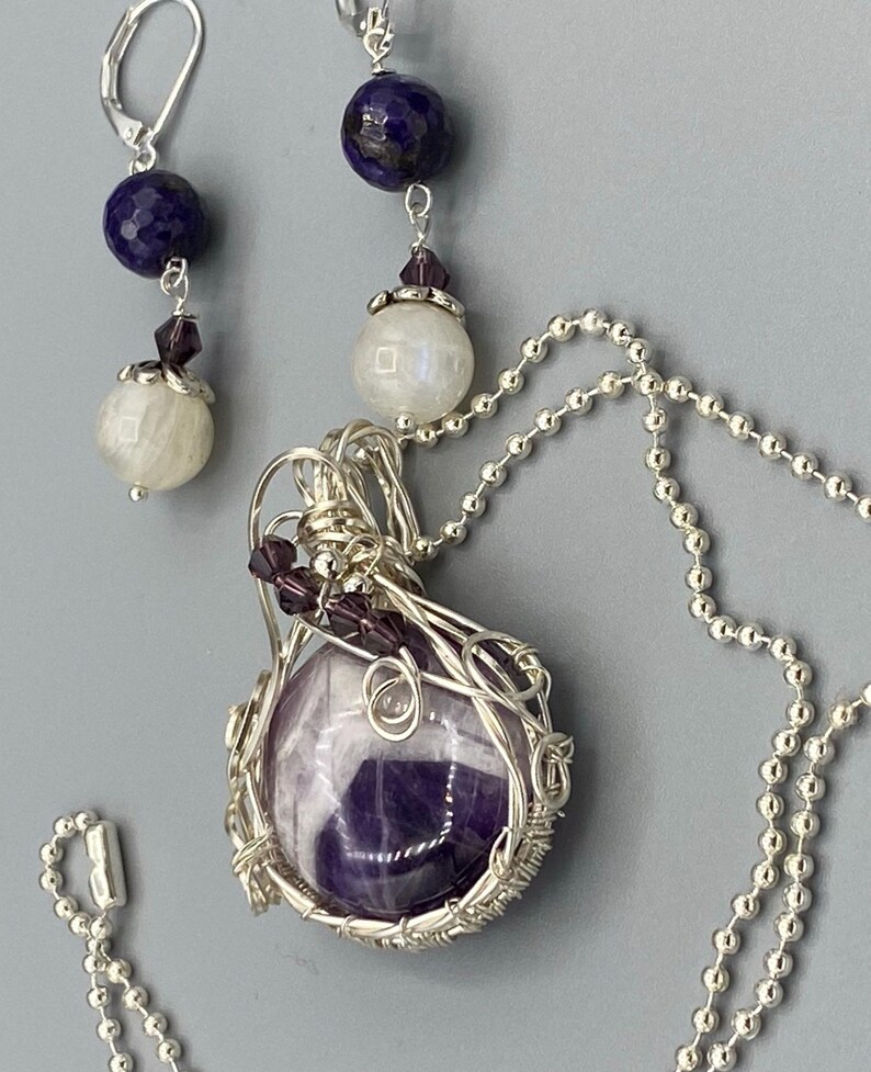 May include: A silver chain necklace with a wire-wrapped amethyst pendant and a silver chain with a pair of earrings featuring a blue stone and a white stone.
