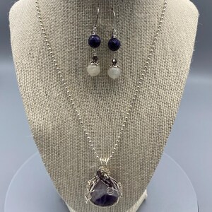 May include: A silver necklace with a purple amethyst stone pendant wrapped in silver wire. The necklace is paired with matching earrings featuring a purple stone and a white pearl.