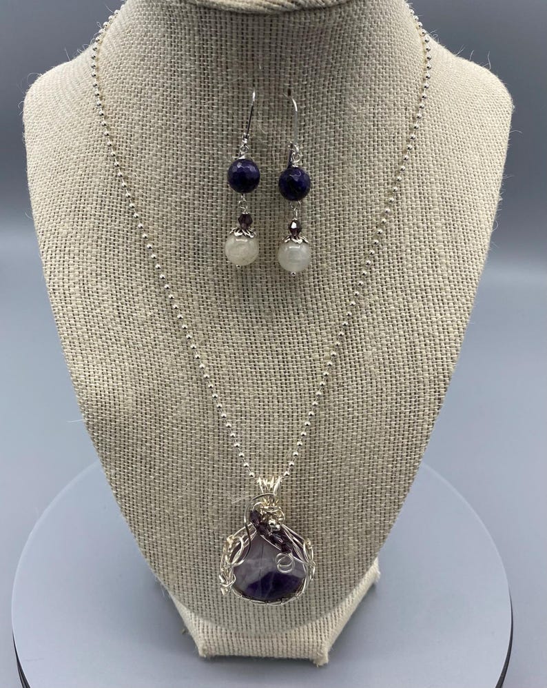 May include: A silver necklace with a purple amethyst stone pendant wrapped in silver wire. The necklace is paired with matching earrings featuring a purple stone and a white pearl.