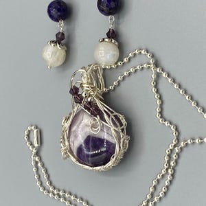 May include: A silver necklace with a purple amethyst stone wrapped in silver wire. The necklace has a silver chain and a silver clasp. The earrings are silver with a purple stone and a white stone.