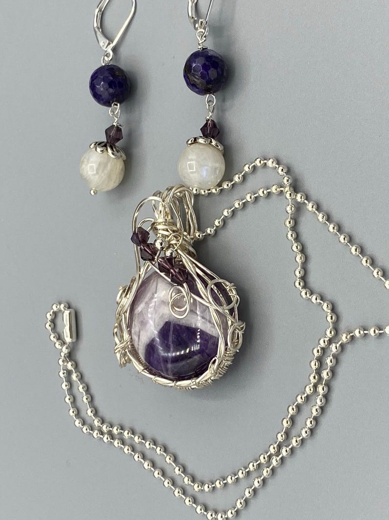 May include: A silver necklace with a purple amethyst stone wrapped in silver wire. The necklace has a silver chain and a silver clasp. The earrings are silver with a purple stone and a white stone.