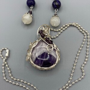 May include: A silver necklace with a purple amethyst stone pendant wrapped in silver wire. The pendant is accented with small purple beads. The necklace is paired with a set of earrings featuring a white moonstone bead and a dark blue lapis lazuli bead.