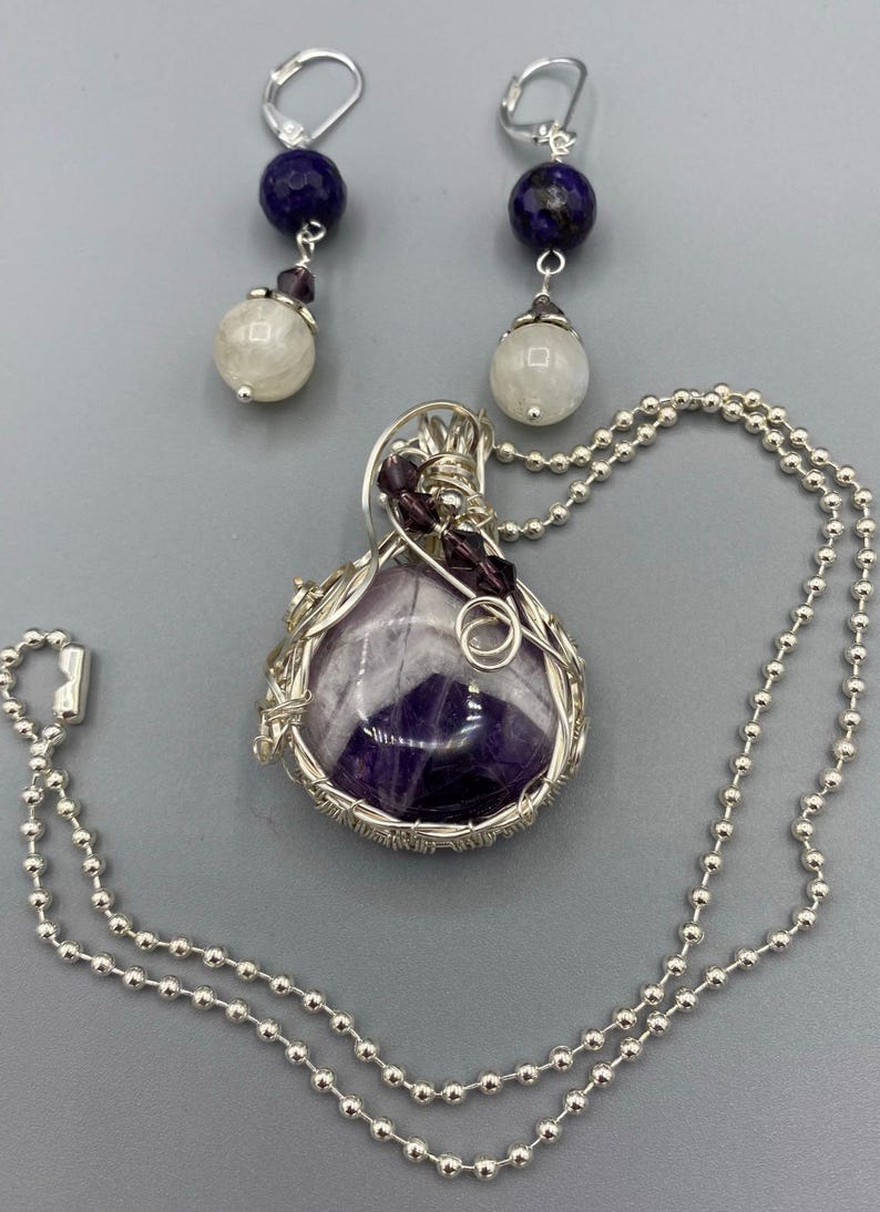 May include: A silver necklace with a purple amethyst stone pendant wrapped in silver wire. The pendant is accented with small purple beads. The necklace is paired with a set of earrings featuring a white moonstone bead and a dark blue lapis lazuli bead.