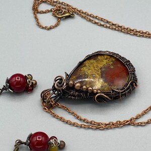May include: A copper wire wrapped pendant necklace with a red, yellow, and gold gemstone. The necklace comes with a pair of matching earrings with red gemstone beads.