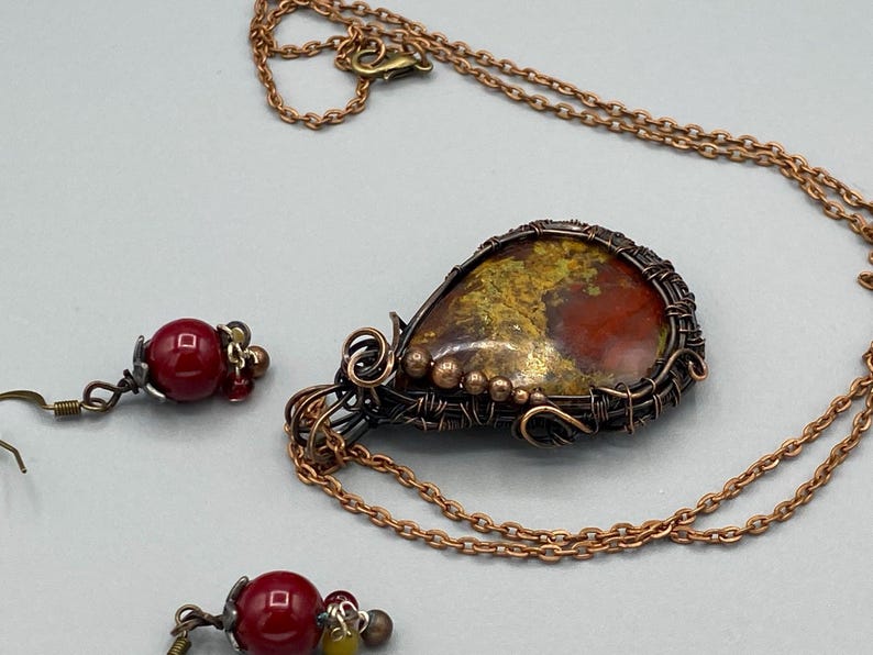 May include: A copper wire wrapped pendant necklace with a red, yellow, and gold gemstone. The necklace comes with a pair of matching earrings with red gemstone beads.