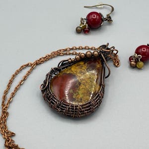 May include: A copper wire wrapped pendant necklace with a red, yellow, and brown stone. The necklace comes with matching earrings with red and gold beads.