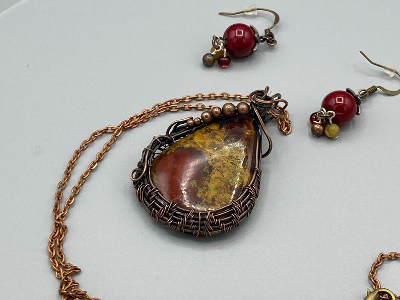 May include: A copper wire wrapped pendant necklace with a red, yellow, and brown stone. The necklace comes with matching earrings with red and gold beads.