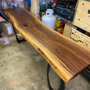 May include: A long, live edge wooden table with a natural wood finish. The table has a unique, organic shape and is supported by two black metal legs. The table is made of walnut wood and has a natural, rustic look.