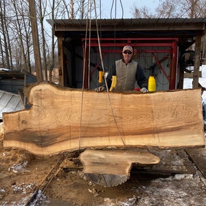 May include: A large slab of wood, likely ash, with a natural edge and a rough surface. The wood is being lifted by a crane and has measurements marked on it: 50", 40", 3", 34", 12 7/4". The wood is labeled "ASH-003710".