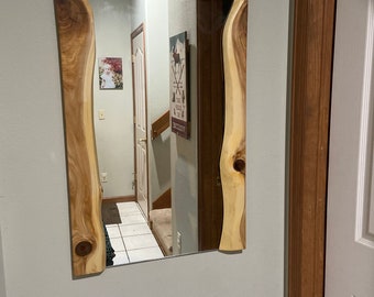 May include: A rectangular mirror with a natural wood frame. The frame has a live edge design with a light brown color and a knot in the wood. The mirror is hanging on a white wall.