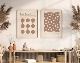 Può includere: Due stampe incorniciate con disegni geometrici in marrone su uno sfondo crema. Le stampe sono intitolate "Bauhaus" e "Exhibition" e sono appese a un muro bianco. Uno scaffale in legno con un cesto intrecciato e libri si trova sotto le stampe. Due sedie in vimini con cuscini bianchi sono su entrambi i lati dello scaffale.