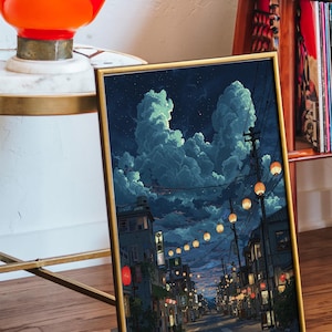 May include: Framed art print depicting a nighttime street scene with glowing lanterns and buildings. The sky is filled with large, fluffy clouds. The artwork has a gold frame and is displayed on a wooden surface.