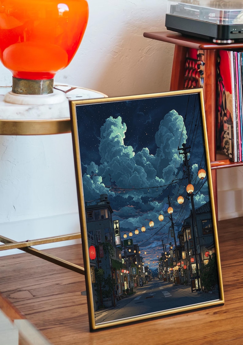 May include: Framed art print depicting a nighttime street scene with glowing lanterns and buildings. The sky is filled with large, fluffy clouds. The artwork has a gold frame and is displayed on a wooden surface.