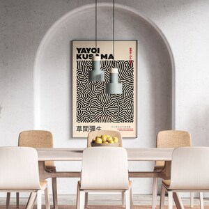 May include: A dining room with a white wall and an arched doorway. A poster with black and white abstract art hangs on the wall. The poster features the text "YAYOI KUSAMA" and Japanese characters. There are four chairs and a table with a bowl of lemons in the center.
