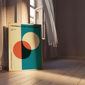 May include: A framed poster with a geometric design featuring overlapping circles in shades of red, yellow, and green on a teal background. The poster has the text "Führend in der Herrenmode" and "Juli-Sept 1925" in black.