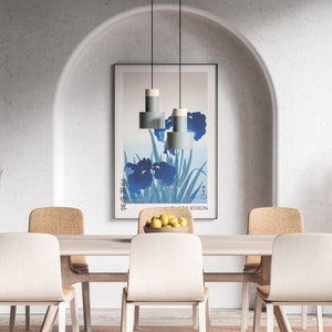 May include: A dining room with a light wood table and six chairs. The table is set with a bowl of lemons and a vase of dried pampas grass. A framed print of blue irises hangs on the wall behind the table. Three light blue pendant lights hang above the table.