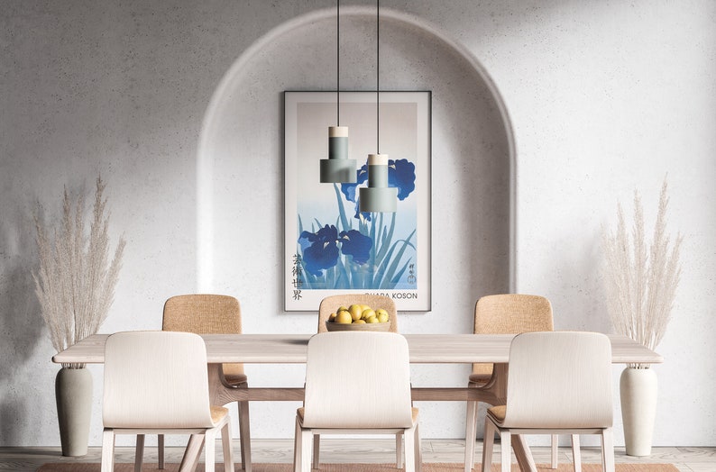 May include: A dining room with a light wood table and six chairs. The table is set with a bowl of lemons and a vase of dried pampas grass. A framed print of blue irises hangs on the wall behind the table. Three light blue pendant lights hang above the table.