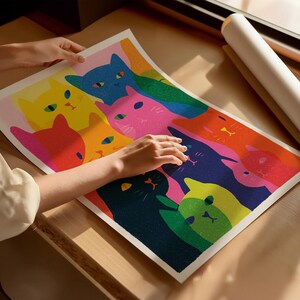 May include: A colorful art print featuring a vibrant illustration of stylized cats in various colors, including pink, yellow, blue, and green. The artwork is held by a person, showcasing the print's size and detail. The print is on a light-colored surface.