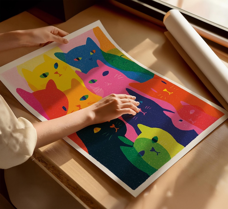 May include: A colorful art print featuring a vibrant illustration of stylized cats in various colors, including pink, yellow, blue, and green. The artwork is held by a person, showcasing the print's size and detail. The print is on a light-colored surface.
