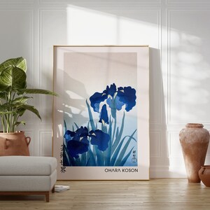 May include: A framed print of a Japanese-style painting of blue irises with green leaves. The print is titled "Ohara Koson" and is displayed on a white wall in a room with a potted plant, a white ottoman, and two terracotta vases.