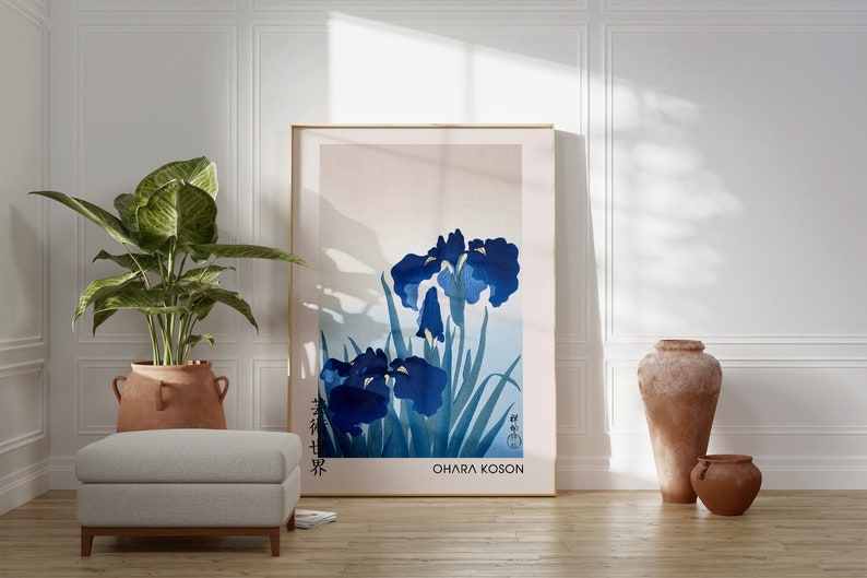 May include: A framed print of a Japanese-style painting of blue irises with green leaves. The print is titled "Ohara Koson" and is displayed on a white wall in a room with a potted plant, a white ottoman, and two terracotta vases.