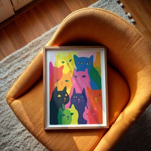 May include: A framed art print featuring a colorful illustration of numerous cats. The cats are depicted in various colors, including pink, blue, green, and orange. The artwork is set against a light pink background and framed in a light wood frame. The print is resting on an orange chair.