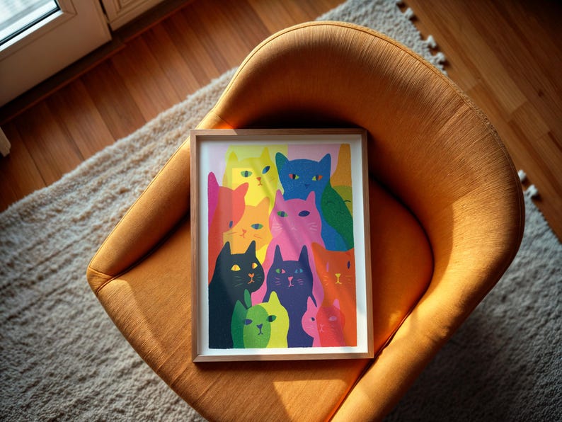 May include: A framed art print featuring a colorful illustration of numerous cats. The cats are depicted in various colors, including pink, blue, green, and orange. The artwork is set against a light pink background and framed in a light wood frame. The print is resting on an orange chair.