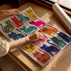 K&ouml;nnte beinhalten: Ein farbenfroher Kunstdruck mit einem sich wiederholenden Muster stilisierter Katzen in verschiedenen Farben, darunter Gelb, Rot, Blau und Pink. Der Druck wird auf einer Holzoberfl&auml;che pr&auml;sentiert, mit einer aufgerollten Posterrolle im Hintergrund.