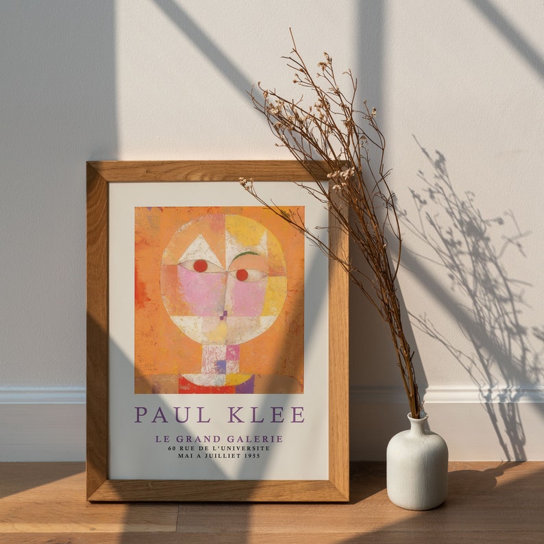 May include: A framed print of a painting by Paul Klee titled "Le Grand Galerie". The painting features a stylized human face with a yellow, pink, and white color scheme. The frame is made of wood and has a light brown finish. The print is displayed on a white wall with a vase of dried flowers in the foreground.