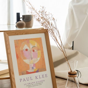 May include: A framed print of a Paul Klee painting titled "Le Grand Galerie" with a geometric abstract design in shades of orange, yellow, pink, and white. The print is in a wooden frame and is displayed on a table with a vase of dried flowers.