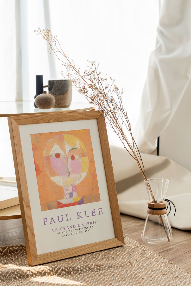 May include: A framed print of a Paul Klee painting titled "Le Grand Galerie" with a geometric abstract design in shades of orange, yellow, pink, and white. The print is in a wooden frame and is displayed on a table with a vase of dried flowers.