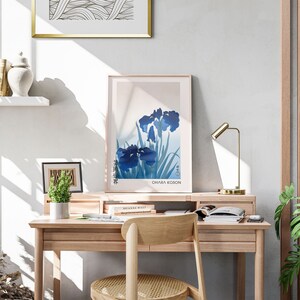May include: A light wood desk with a gold lamp, a framed print of blue irises with the text "Ohara Koson", and a potted plant. The desk has a chair with a woven seat.