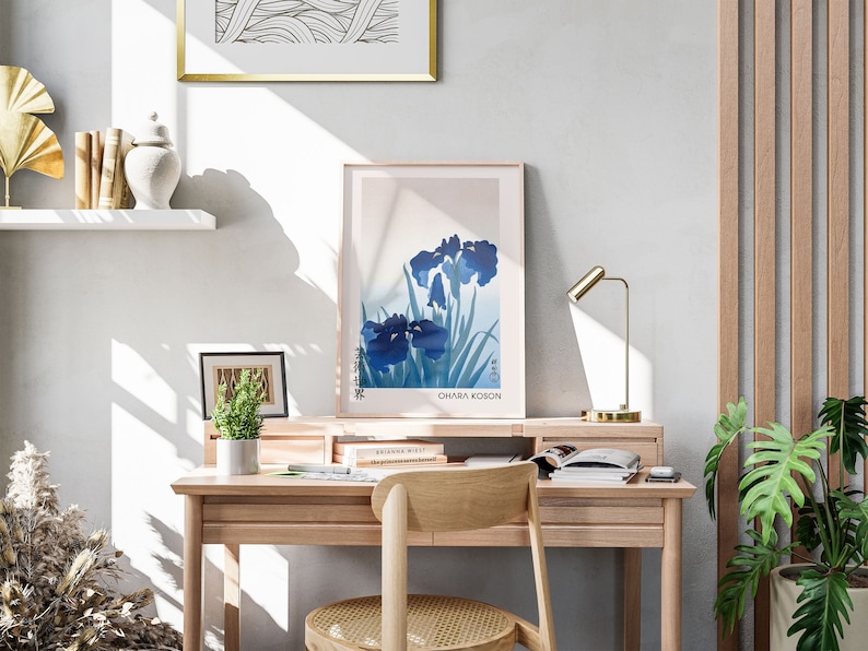 May include: A light wood desk with a gold lamp, a framed print of blue irises with the text "Ohara Koson", and a potted plant. The desk has a chair with a woven seat.