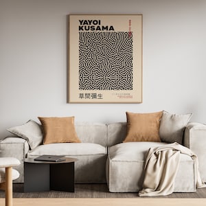 May include: A living room interior with a white couch, two brown throw pillows, a beige throw blanket, a black side table, a black floor lamp, and a framed poster with black and white abstract art. The poster has the text "Yayoi Kusama" and Japanese characters.