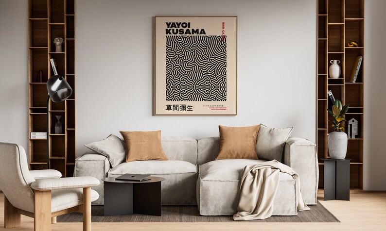 May include: A living room interior with a white couch, two brown throw pillows, a beige throw blanket, a black side table, a black floor lamp, and a framed poster with black and white abstract art. The poster has the text "Yayoi Kusama" and Japanese characters.