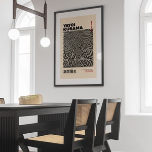 May include: A black framed poster with a white background featuring a black and white abstract design. The poster has the text "Yayoi Kusama" and "草間彌生" in black text. The poster is hanging on a white wall above a black dining table with two black chairs.