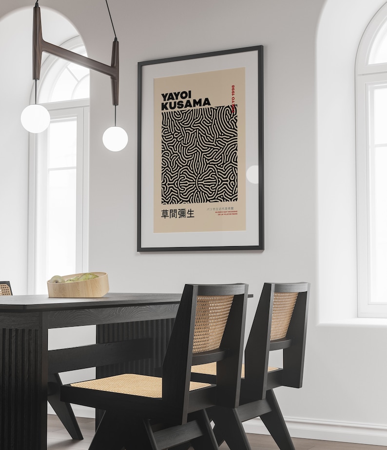 May include: A black framed poster with a white background featuring a black and white abstract design. The poster has the text "Yayoi Kusama" and "草間彌生" in black text. The poster is hanging on a white wall above a black dining table with two black chairs.