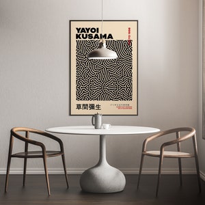 May include: A black and white poster with a geometric pattern and the text "YAYOI KUSAMA 1929-." and "草間彌生" in Japanese. The poster is hanging on a white wall above a white table with two wooden chairs.