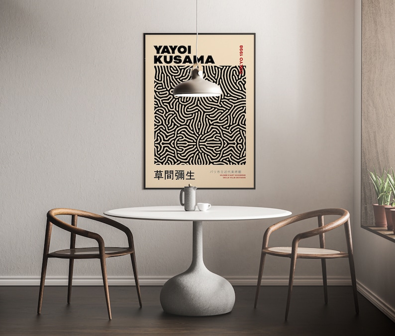 May include: A black and white poster with a geometric pattern and the text "YAYOI KUSAMA 1929-." and "草間彌生" in Japanese. The poster is hanging on a white wall above a white table with two wooden chairs.