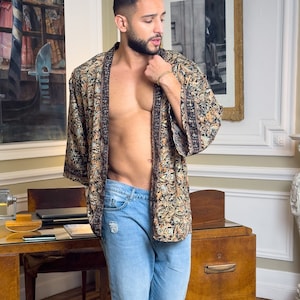 Może przedstawiać: Mężczyzna w brązowo-czarnym kimonie z niebieską dżinsową koszulą pod spodem. Stoi w pokoju z drewnianym biurkiem i dywanem na podłodze.