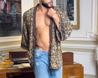May include: A man wearing a brown and black patterned kimono with a blue denim shirt underneath. He is standing in a room with a wooden desk and a rug on the floor.
