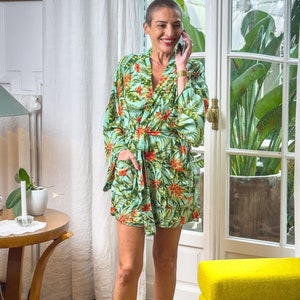 Op de afbeelding: Een vrouw draagt een groene en oranje bloemenprint-badjas met een rode riem. Ze staat in een kamer met een gele bank en een houten vloer.