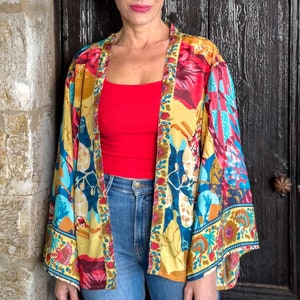 Puede incluir: Una mujer con un kimono floral amarillo, azul y rojo con un top rojo y pantalones vaqueros azules.