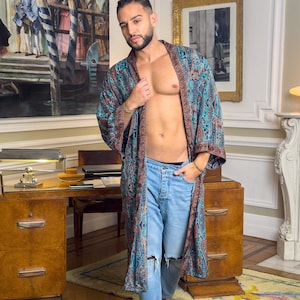 May include: A man wearing a long, blue and brown patterned robe with a paisley design. He is wearing blue jeans with a distressed knee and is barefoot. The robe has a sash that is tied at the waist. The man is standing in a room with a wooden floor and a rug.