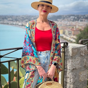 May include: A woman wearing a straw hat, a red tank top, blue jeans, and a colourful floral kimono. She is holding a large, round, woven straw bag.