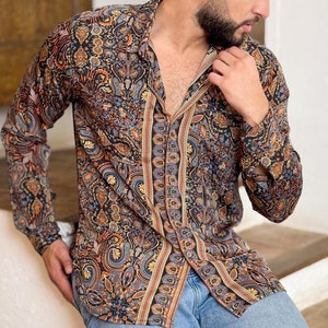 May include: A man wearing a long-sleeved shirt with a paisley pattern in shades of brown, blue, and orange. He is wearing blue jeans with a distressed knee.