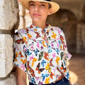 Peut inclure: Un chapeau de paille avec un ruban bleu marine est porté. La personne porte un chemisier blanc orné d'un motif de papillons colorés, un jean bleu et une ceinture marron. Une montre rouge est visible au poignet.